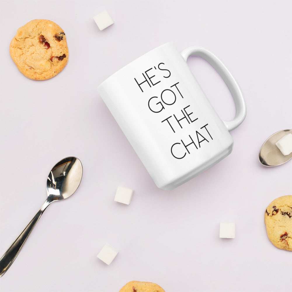 A glossy white ceramic mug with thin, all-caps black sans serif font that reads 'HE'S GOT THE CHAT', lies on its side against a very pale lavender blush background. Surrounding the mug are a few chocolate chip cookies, five cubes of sugar, and two spoons.