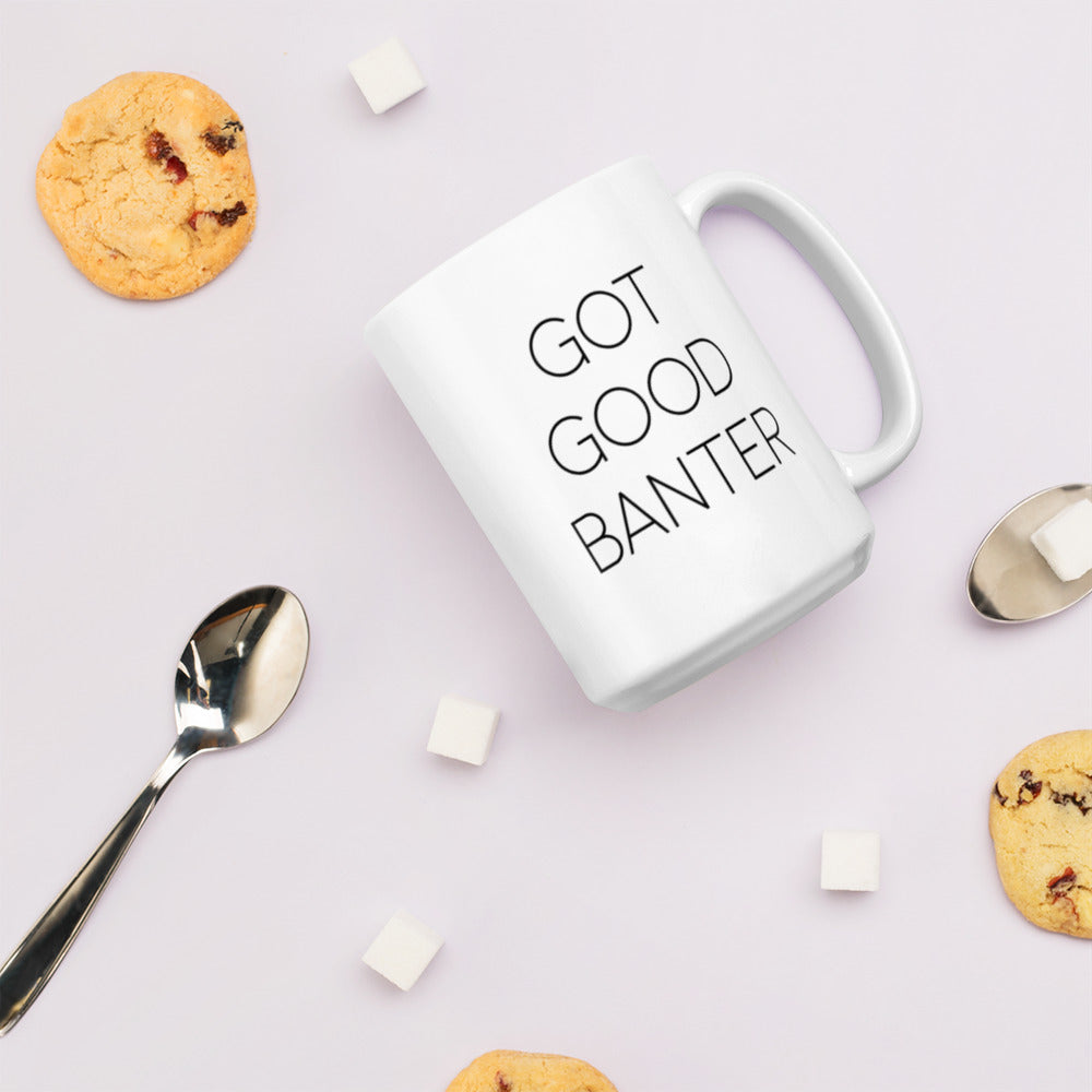 A glossy white ceramic mug with thin, all-caps black sans serif font that reads 'GOT GOOD BANTER', lies on its side against a very pale lavender blush background. Surrounding the mug are a few chocolate chip cookies, five cubes of sugar, and two spoons.