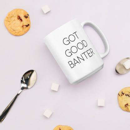 A glossy white ceramic mug with thin, all-caps black sans serif font that reads 'GOT GOOD BANTER', lies on its side against a very pale lavender blush background. Surrounding the mug are a few chocolate chip cookies, five cubes of sugar, and two spoons.