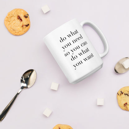 A glossy white ceramic mug with lowercase black serif font that reads 'do what you need so you can do what you want', lies on its side against a very pale lavender blush background. Surrounding the mug are a few chocolate chip cookies, five cubes of sugar, and two spoons.