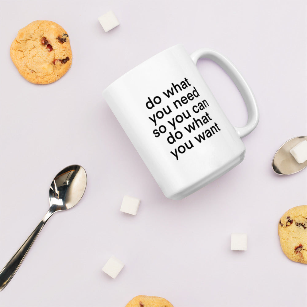 A glossy white ceramic mug with lowercase bolded black sans serif font that reads 'do what you need so you can do what you want', lies on its side against a very pale lavender blush background. Surrounding the mug are a few chocolate chip cookies, five cubes of sugar, and two spoons.