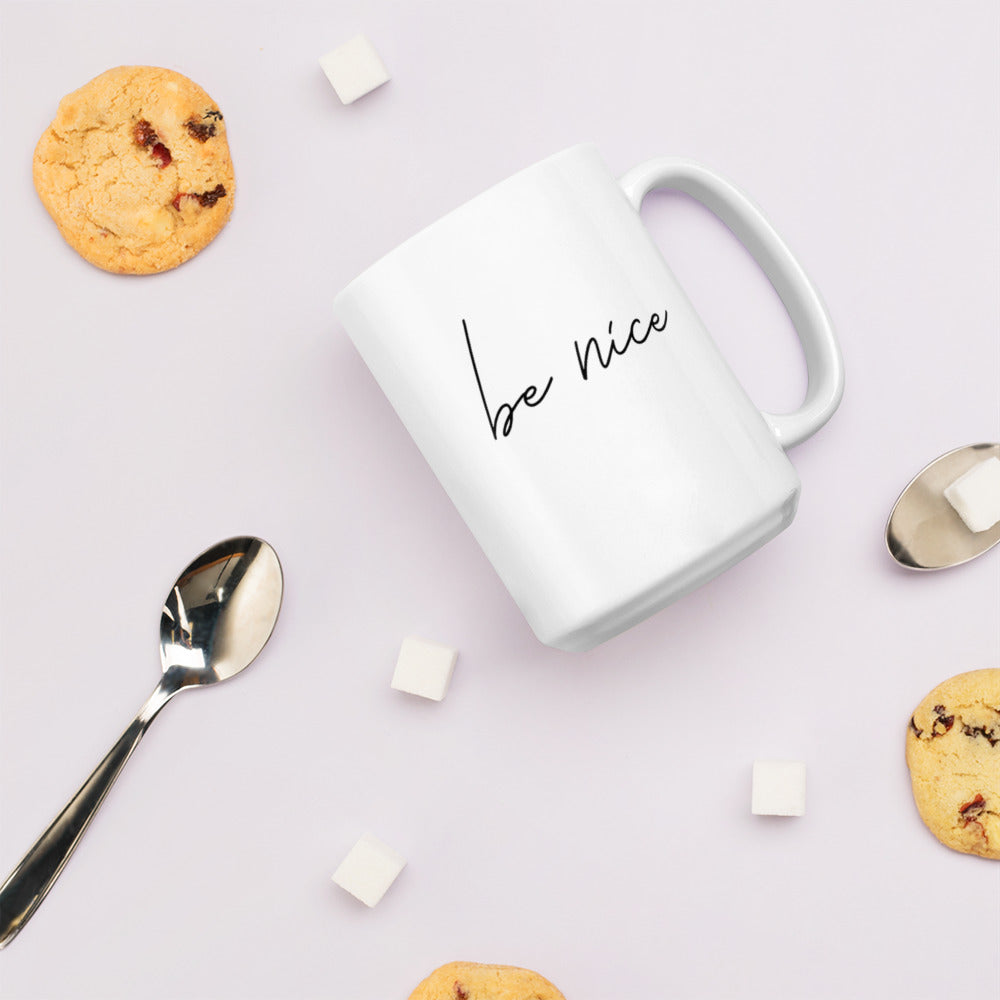 A glossy white ceramic mug with script text that reads 'be nice', lies on its side against a very pale lavender blush background. Surrounding the mug are a few chocolate chip cookies, five cubes of sugar, and two spoons.