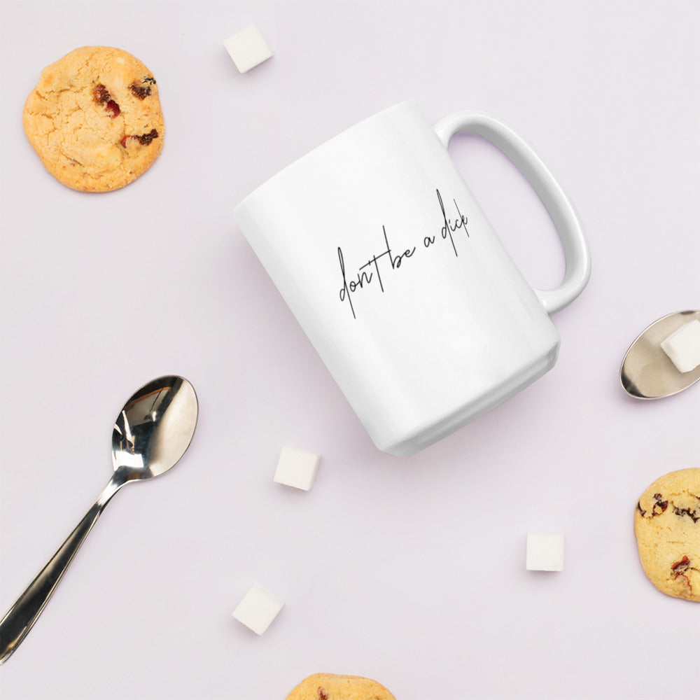 A glossy white ceramic mug with lowercase script font that reads 'don't be a dick', lies on its side against a very pale lavender blush background. Surrounding the mug are a few chocolate chip cookies, five cubes of sugar, and two spoons.