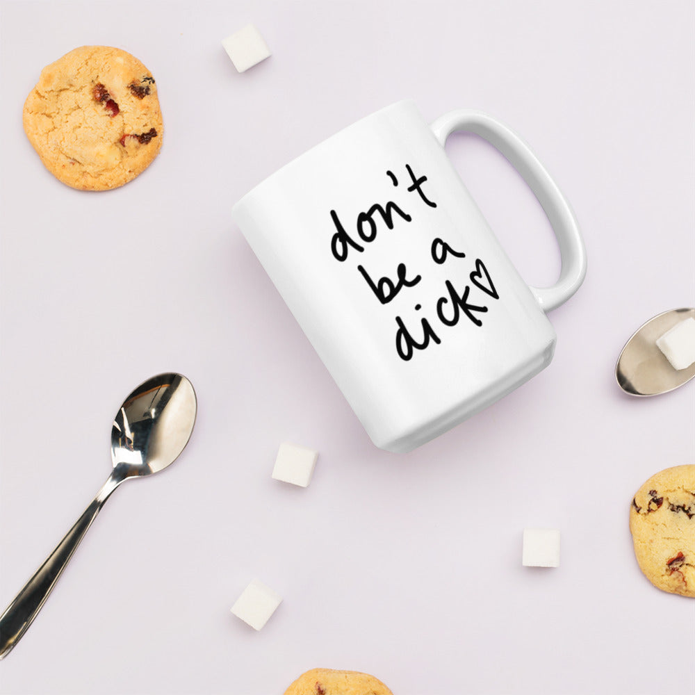 A glossy white ceramic mug with bold handwritten-style font that reads 'don't be a dick [ hand-drawn heart ]', lies on its side against a very pale lavender blush background. Surrounding the mug are a few chocolate chip cookies, five cubes of sugar, and two spoons.