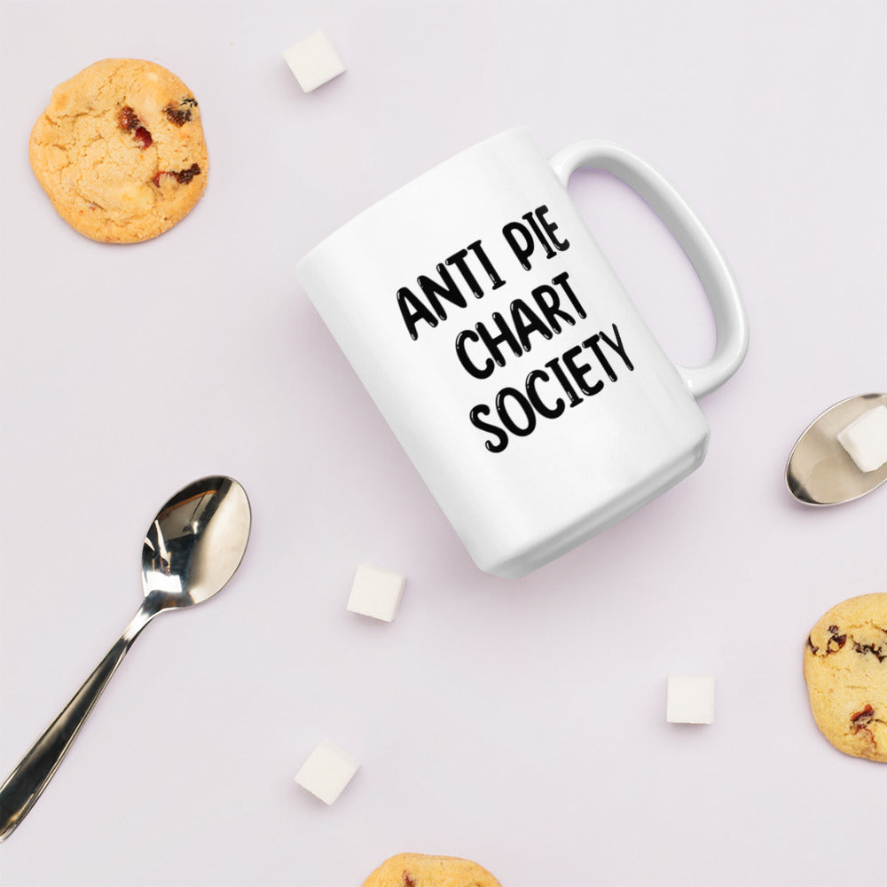 A glossy white ceramic mug with all-uppercase black bubble text that reads 'ANTI PIE CHART SOCIETY', lies on its side against a very pale lavender blush background. Surrounding the mug are a few chocolate chip cookies, five cubes of sugar, and two spoons.