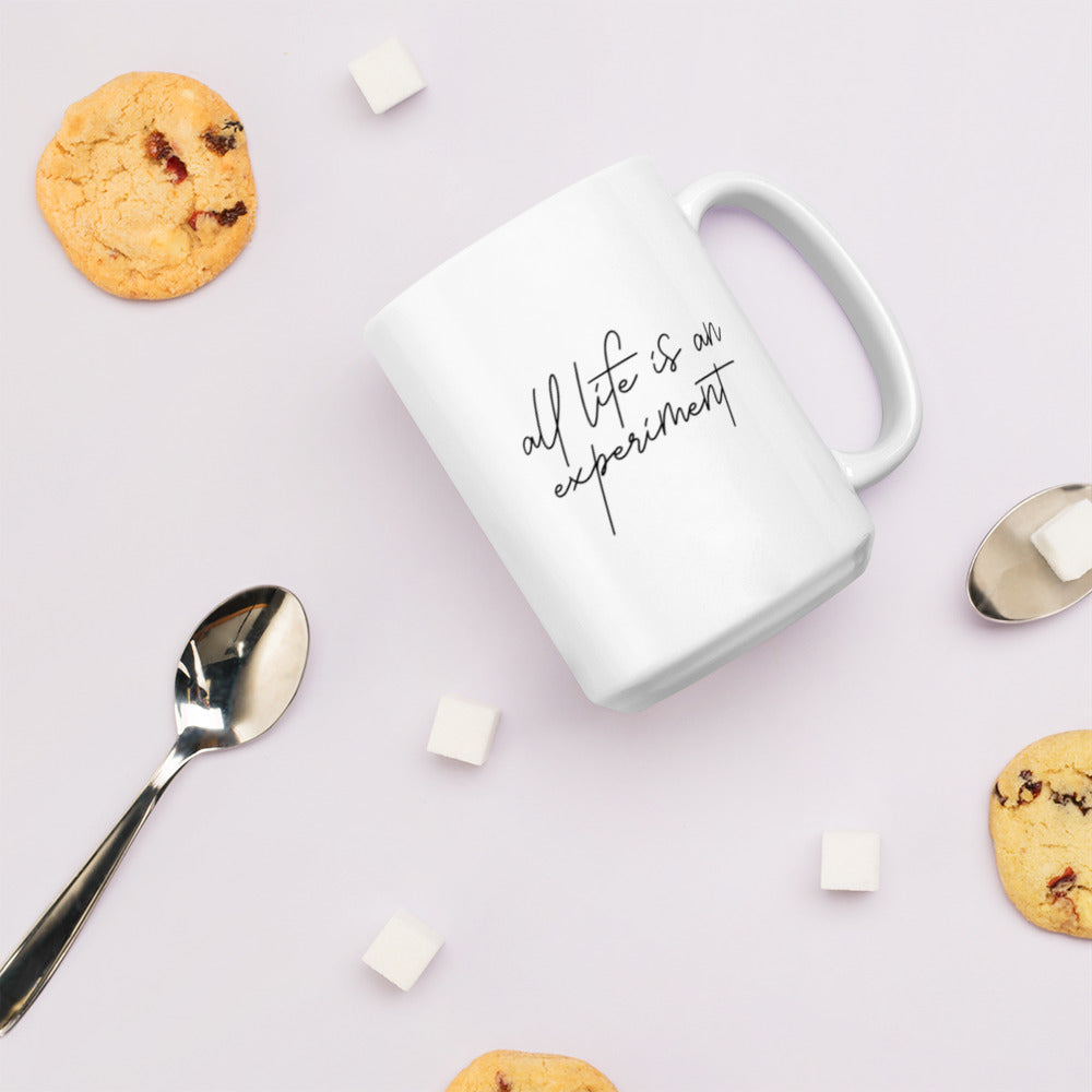 A glossy white ceramic mug with black typography in a script-like font that reads 'all life is an experiment', lies on its side against a very pale lavender blush background. Surrounding the mug are a few chocolate chip cookies, five cubes of sugar, and two spoons.