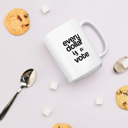 A glossy white ceramic mug with bold, lowercase black sans serif font that reads 'every dollar is a vote', with 'is a' in black handwritten script font, lies on its side against a very pale lavender blush background. Surrounding the mug are a few chocolate chip cookies, five cubes of sugar, and two spoons.