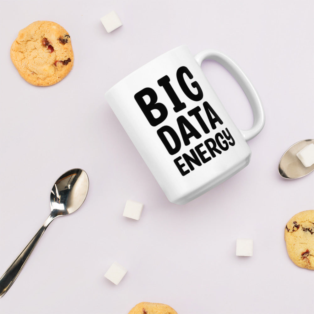 A glossy white ceramic mug with bolded black bubble text that reads 'BIG DATA ENERGY', lies on its side against a very pale lavender blush background. Surrounding the mug are a few chocolate chip cookies, five cubes of sugar, and two spoons.