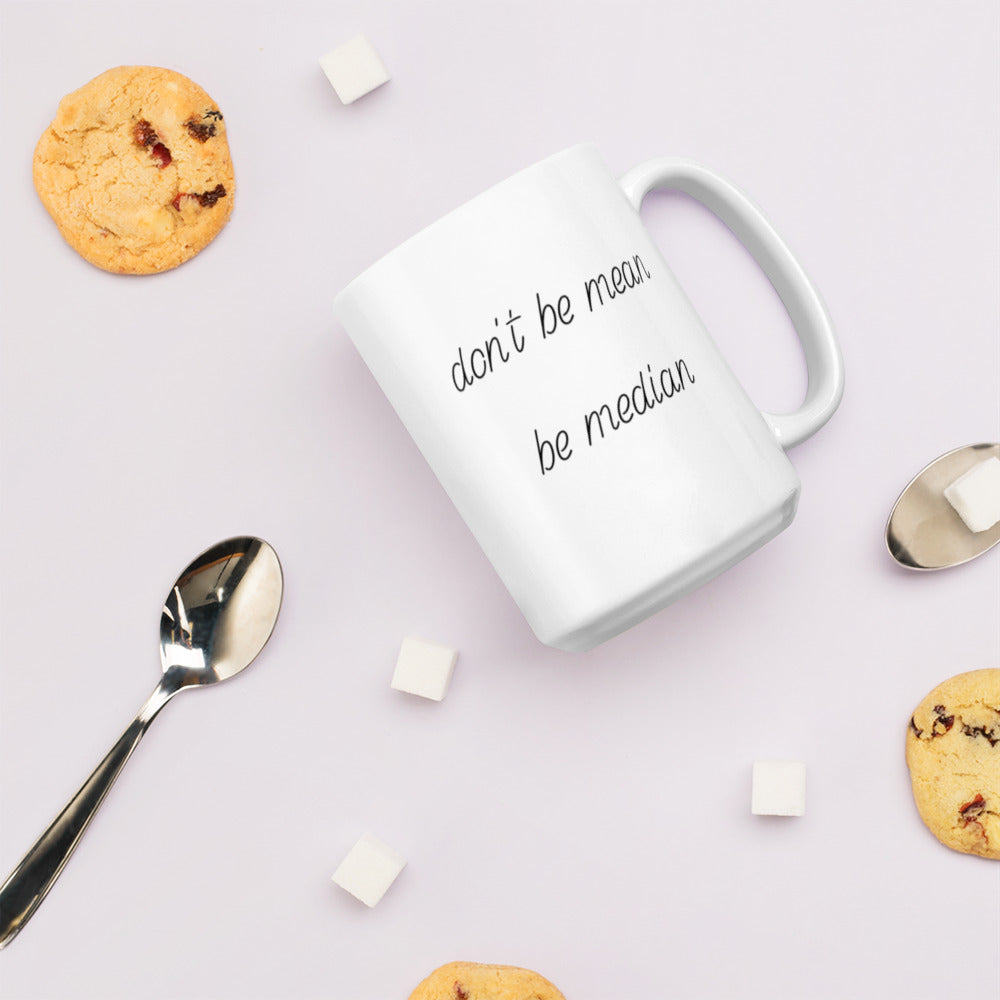 A glossy white ceramic mug with italicized, script-like font that reads 'don't be mean be median', lies on its side against a very pale lavender blush background. Surrounding the mug are a few chocolate chip cookies, five cubes of sugar, and two spoons.