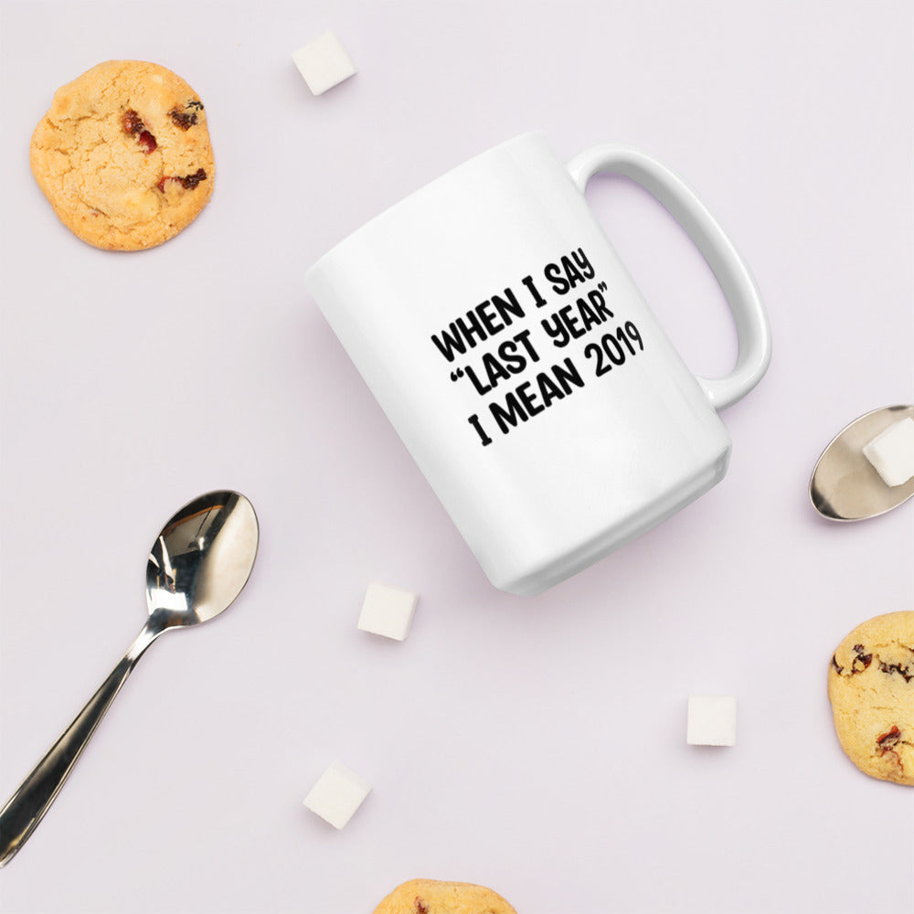 A glossy white ceramic mug with the text 'WHEN I SAY 'LAST YEAR' I MEAN 2019' in black bold uppercase font, lies on its side against a very pale lavender blush background. Surrounding the mug are a few chocolate chip cookies, five cubes of sugar, and two spoons.