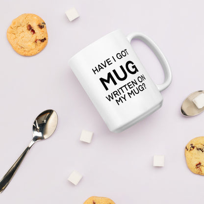 A glossy white ceramic mug with black bolded sans serif text that reads 'HAVE I GOT MUG WRITTEN ON MY MUG?', where the first 'MUG' is in bigger font size, lies on its side against a very pale lavender blush background. Surrounding the mug are a few chocolate chip cookies, five cubes of sugar, and two spoons.