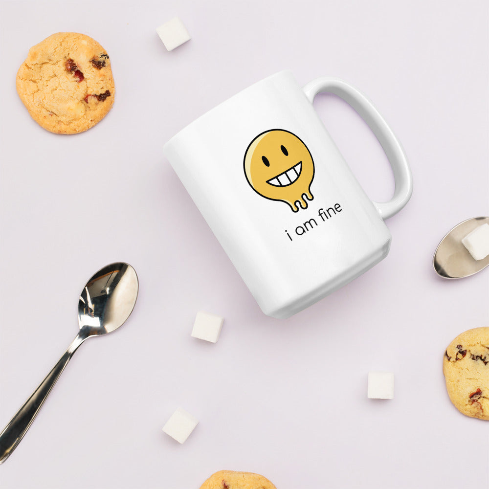 A glossy white ceramic mug with a graphic of a yellow smiley face (grinning) and melting at the bottom, accompanied by the caption 'i am fine' in small black sans serif text, lies on its side against a very pale lavender blush background. Surrounding the mug are a few chocolate chip cookies, five cubes of sugar, and two spoons.