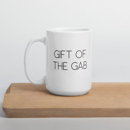 A glossy white ceramic mug with thin, all-caps black sans serif font that reads 'GIFT OF THE GAB', positioned on a wooden cutting board against a neutral background.