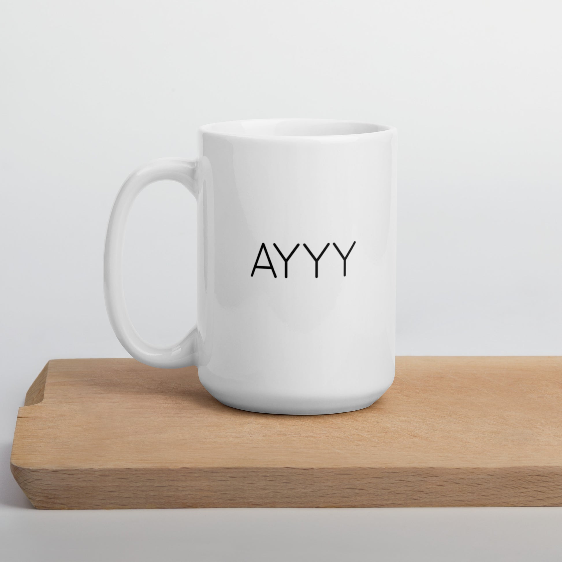 A glossy white ceramic mug with all-caps thin black text that reads 'AYYY', positioned on a wooden cutting board against a neutral background.