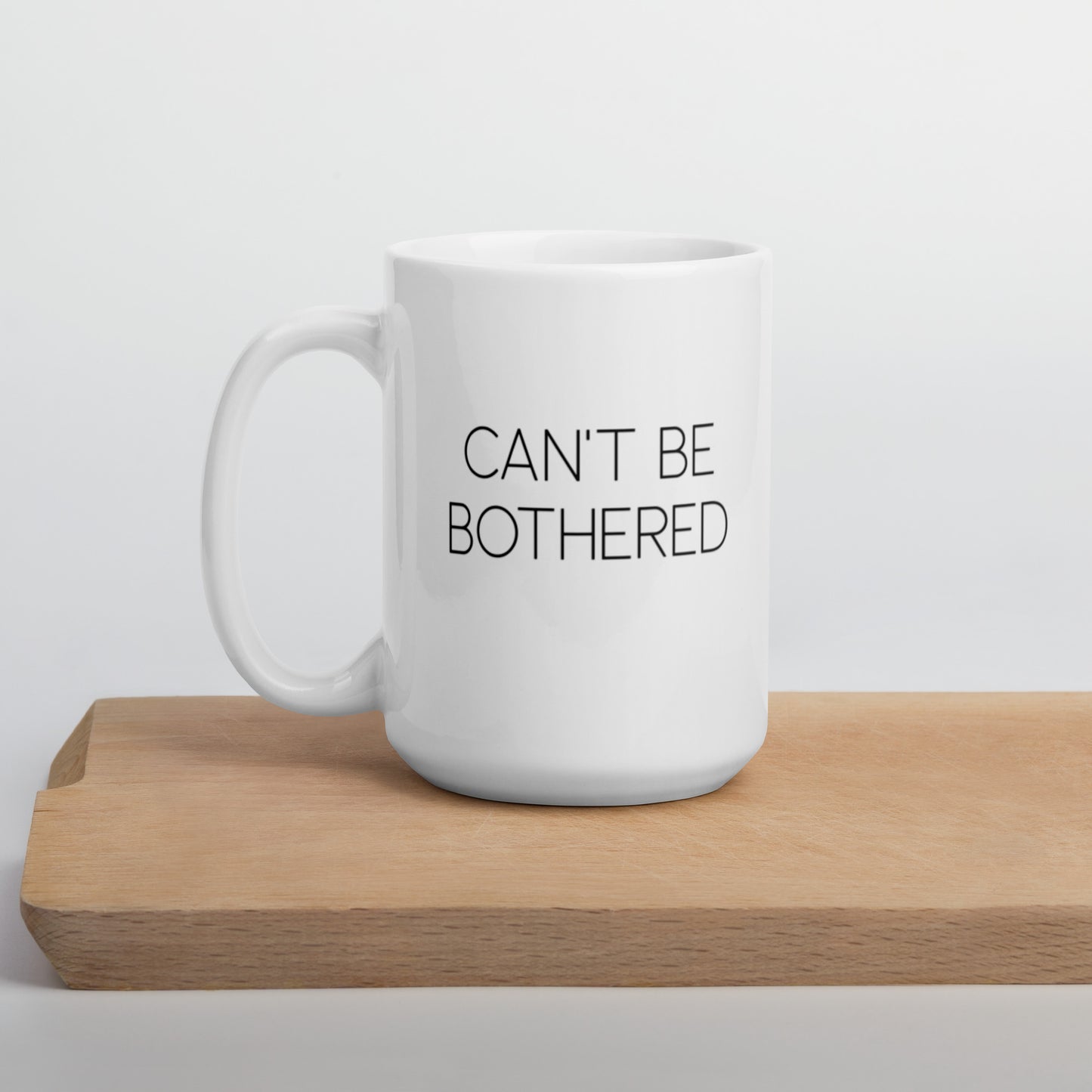 A glossy white ceramic mug with thin uppercase black text that reads 'CAN'T BE BOTHERED', positioned on a wooden cutting board against a neutral background.