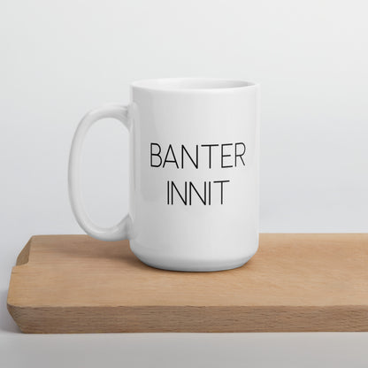 A glossy white ceramic mug with all-caps thin black text that reads 'BANTER INNIT', positioned on a wooden cutting board against a neutral background.