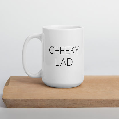 A glossy white ceramic mug with thin black all-caps text that reads 'CHEEKY LAD', positioned on a wooden cutting board against a neutral background.