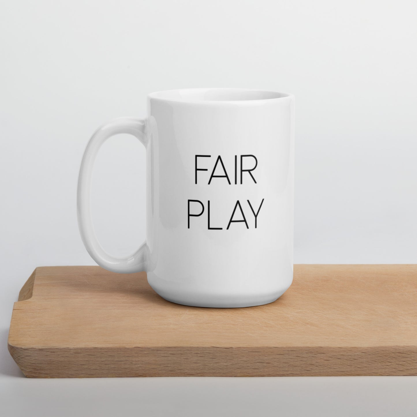 A glossy white ceramic mug with thin, all-caps black sans serif font that reads 'FAIR PLAY', positioned on a wooden cutting board against a neutral background.