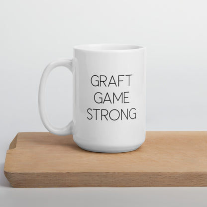 A glossy white ceramic mug with thin, all-caps black sans serif font that reads 'GRAFT GAME STRONG', positioned on a wooden cutting board against a neutral background.