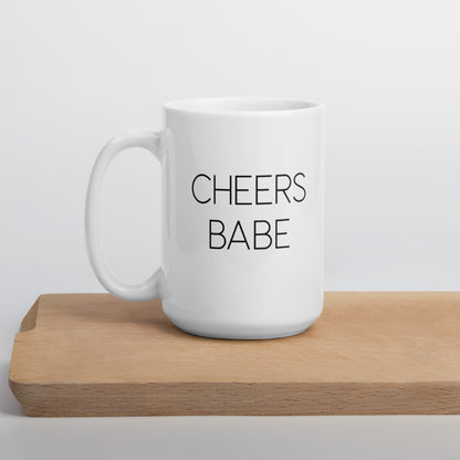 A glossy white ceramic mug with thin black all-caps text that reads 'CHEERS BABE', positioned on a wooden cutting board against a neutral background.