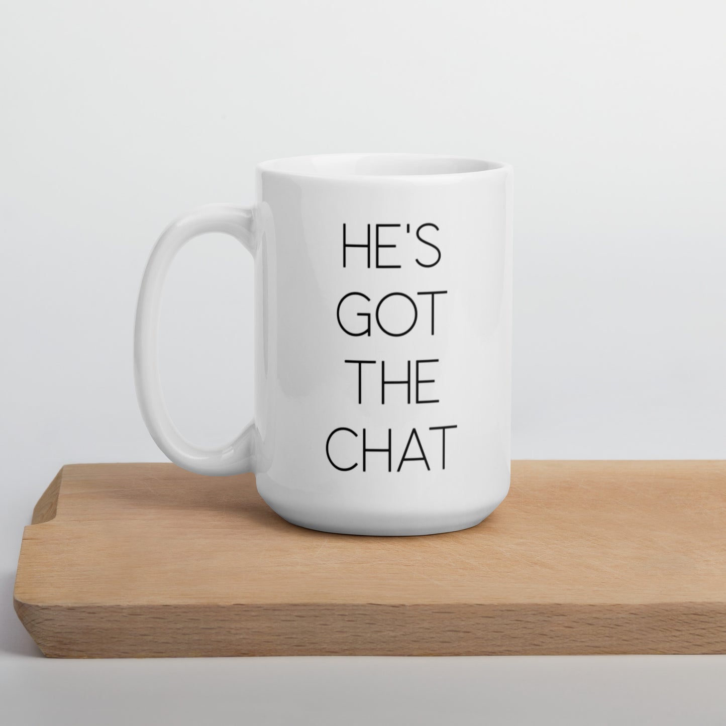 A glossy white ceramic mug with thin, all-caps black sans serif font that reads 'HE'S GOT THE CHAT', positioned on a wooden cutting board against a neutral background.