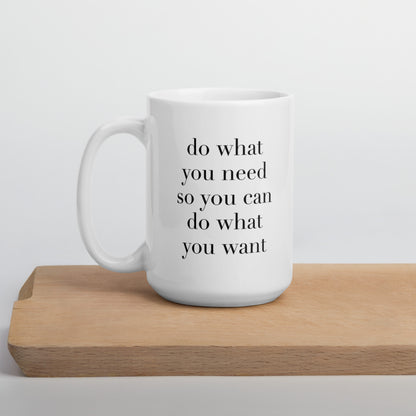 A glossy white ceramic mug with lowercase black serif font that reads 'do what you need so you can do what you want', positioned on a wooden cutting board against a neutral background.