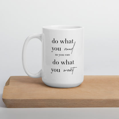 A glossy white ceramic mug with lowercase black serif font that reads 'do what you need so you can do what you want' ('so you can' is in smaller font, and 'need' and 'want' are in script to provide contrast with the serif font), positioned on a wooden cutting board against a neutral background.