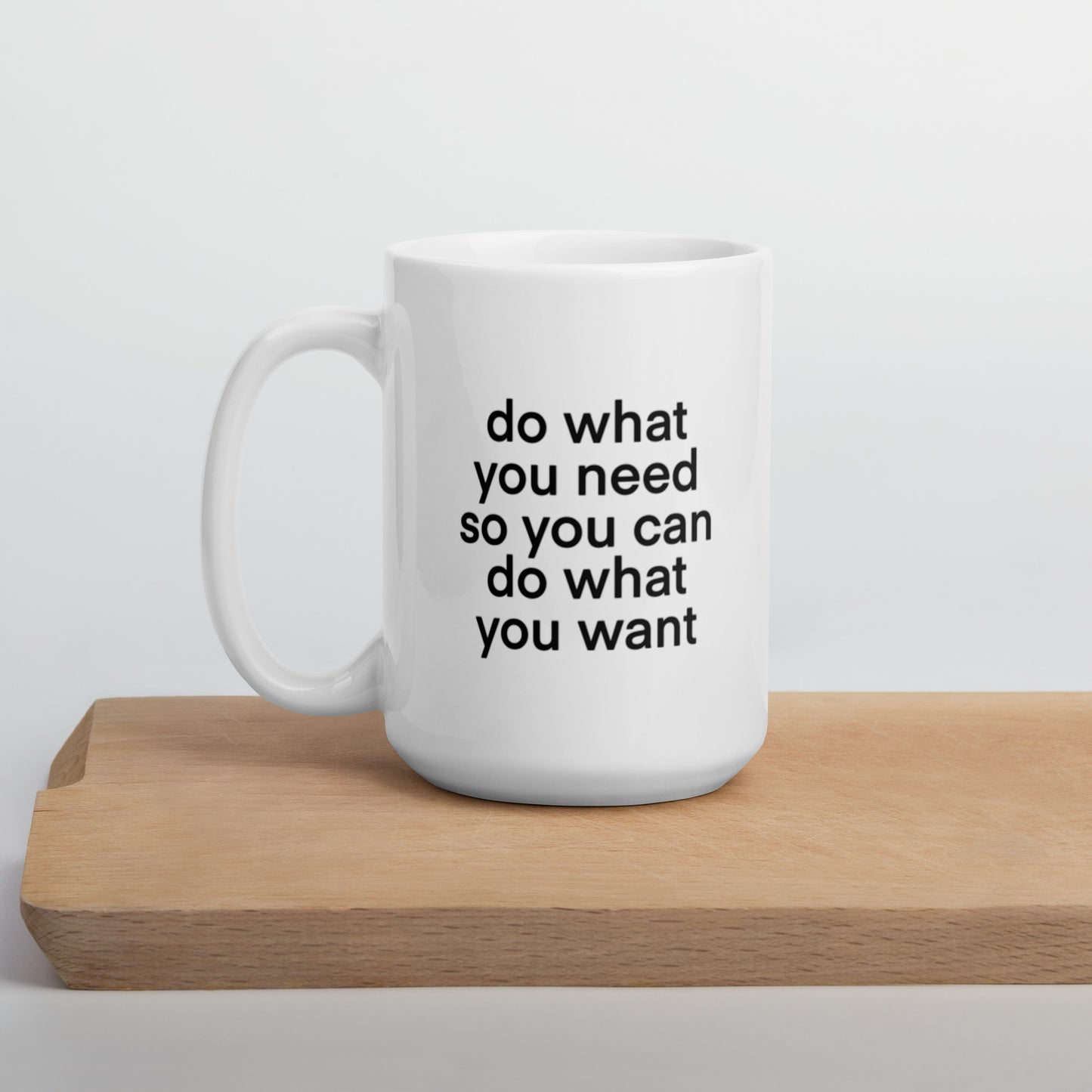 A glossy white ceramic mug with lowercase bolded black sans serif font that reads 'do what you need so you can do what you want', positioned on a wooden cutting board against a neutral background.