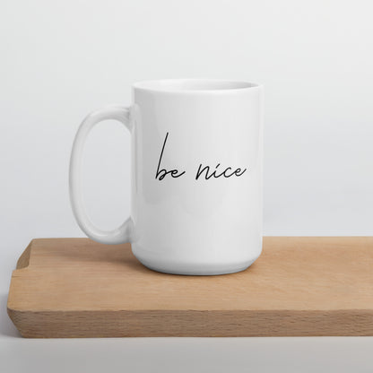 A glossy white ceramic mug with black script text that reads 'be nice', positioned on a wooden cutting board against a neutral background.