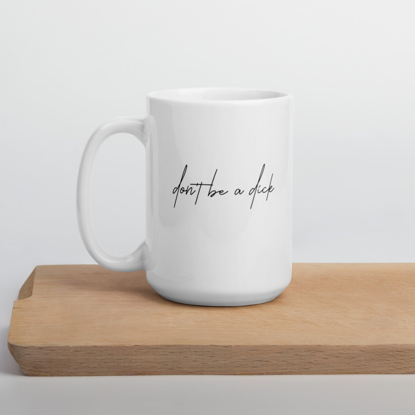 A glossy white ceramic mug with lowercase script font that reads 'don't be a dick', positioned on a wooden cutting board against a neutral background.