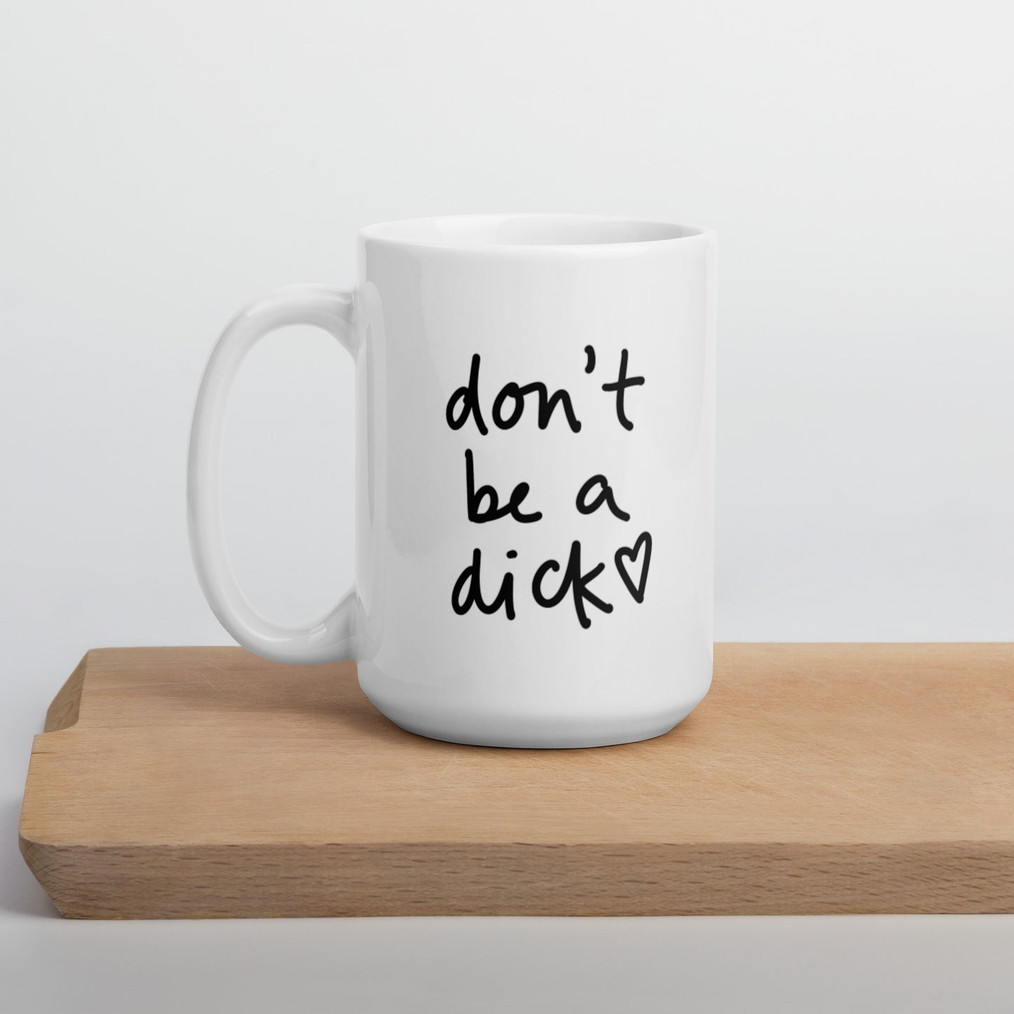 A glossy white ceramic mug with bold handwritten-style font that reads 'don't be a dick [ hand-drawn heart ]', positioned on a wooden cutting board against a neutral background.