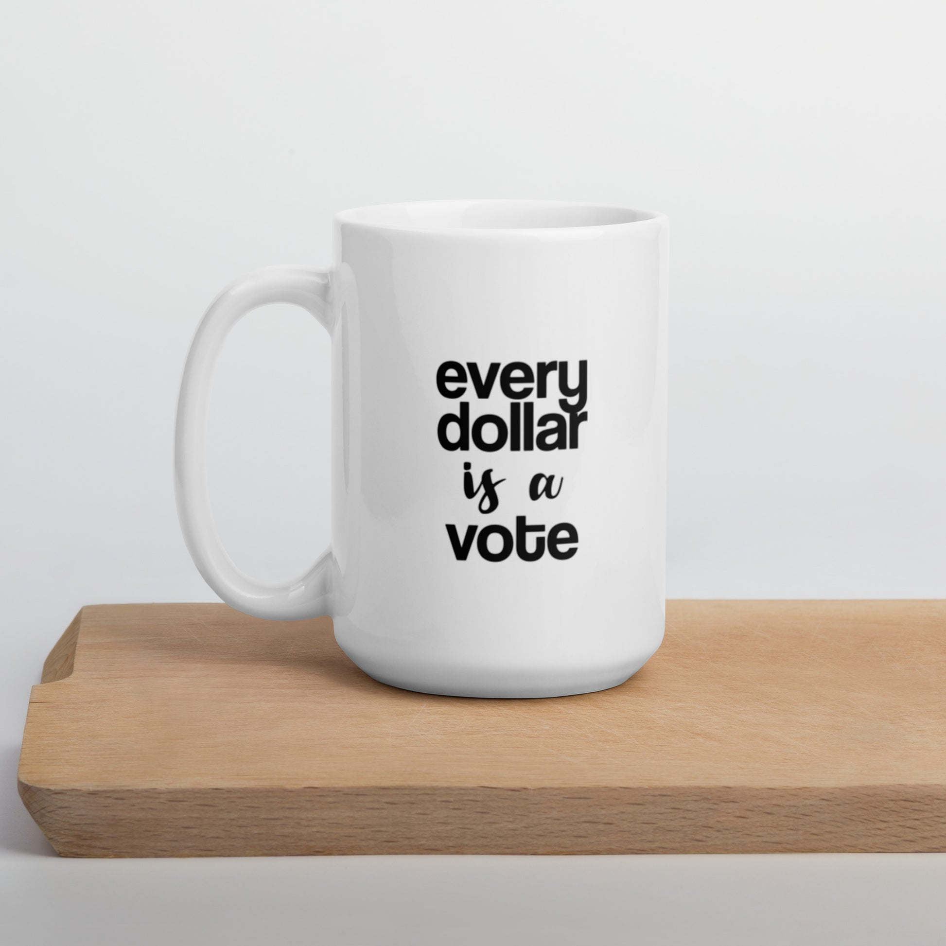 A glossy white ceramic mug with bold, lowercase black sans serif font that reads 'every dollar is a vote', with 'is a' in black handwritten script font, positioned on a wooden cutting board against a neutral background.