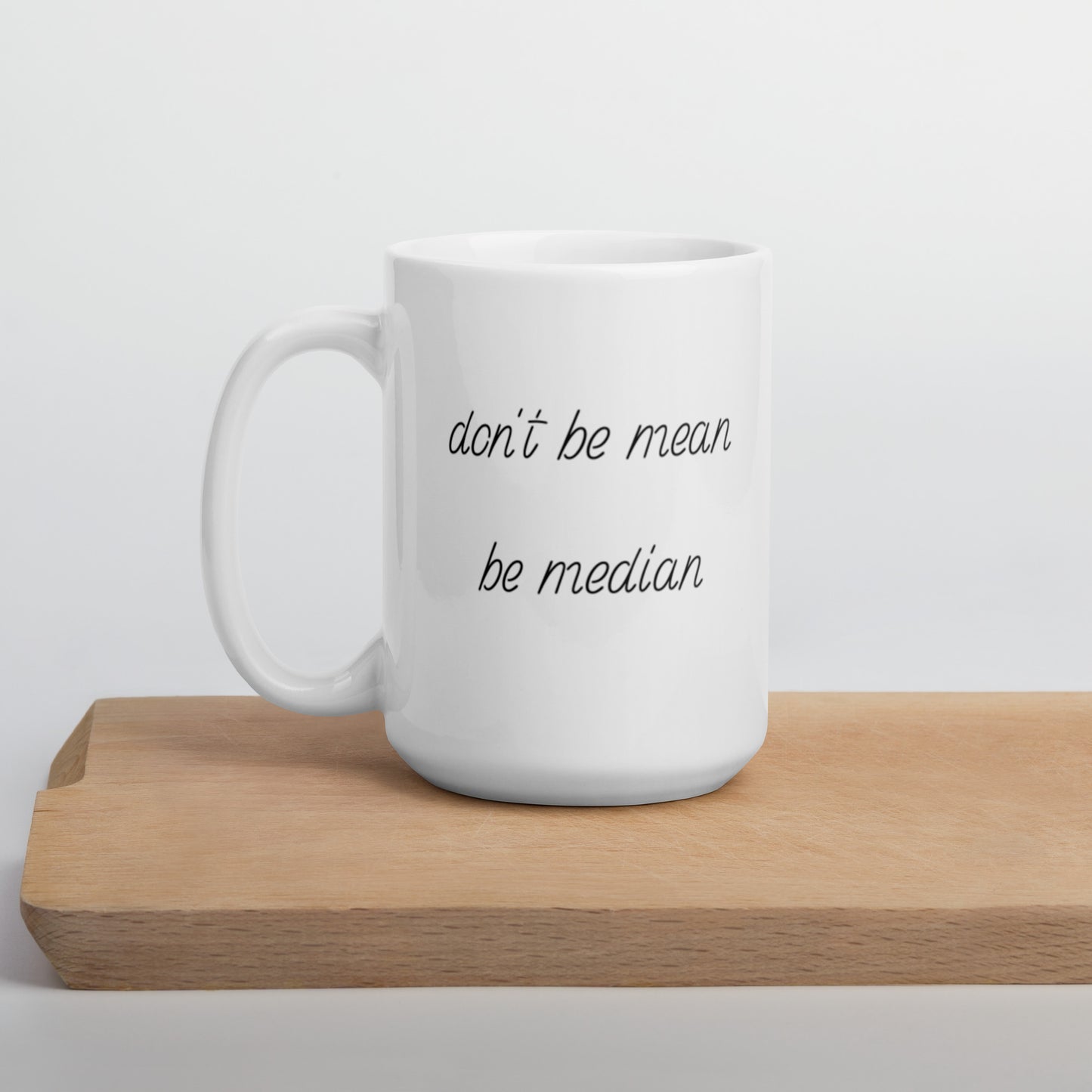 A glossy white ceramic mug with italicized, script-like font that reads 'don't be mean be median', positioned on a wooden cutting board against a neutral background.