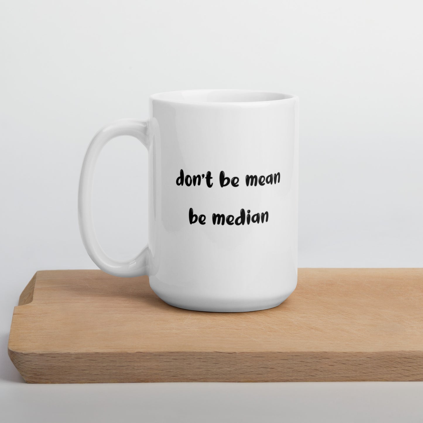 A glossy white ceramic mug with bold, lowercase black bubble font that reads 'don't be mean be median', positioned on a wooden cutting board against a neutral background.