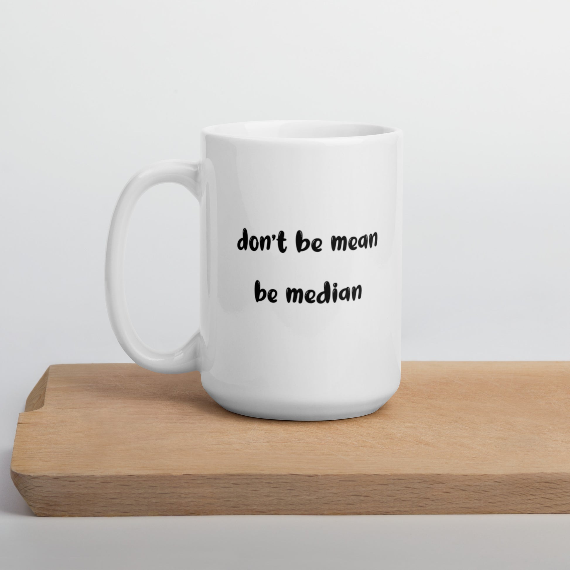 A glossy white ceramic mug with bold, lowercase black bubble font that reads 'don't be mean be median', positioned on a wooden cutting board against a neutral background.