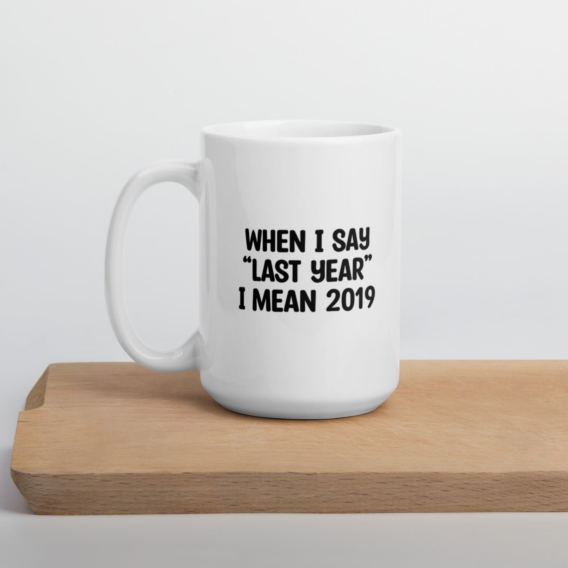 A glossy white ceramic mug with the text 'WHEN I SAY 'LAST YEAR' I MEAN 2019' in black bold uppercase font, positioned on a wooden cutting board against a neutral background.