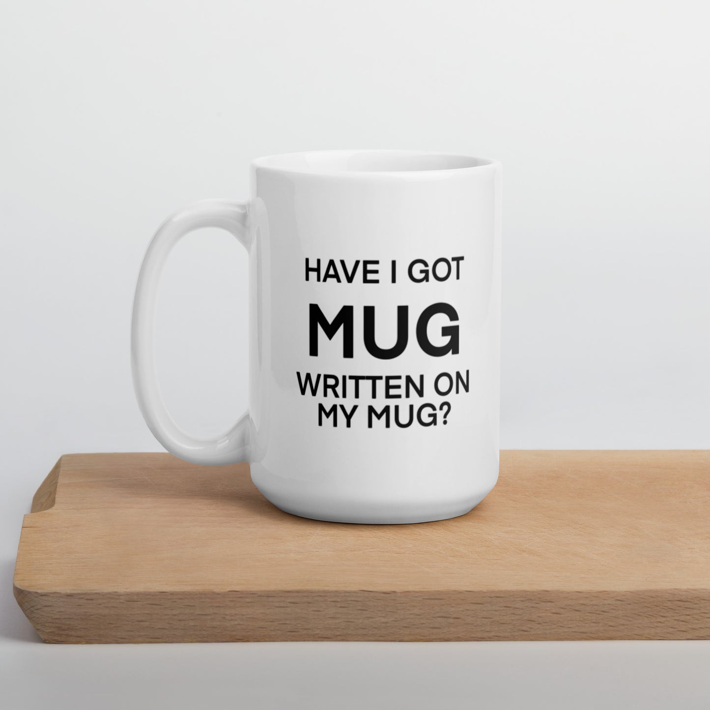 A glossy white ceramic mug with black bolded sans serif text that reads 'HAVE I GOT MUG WRITTEN ON MY MUG?', where the first 'MUG' is in bigger font size, positioned on a wooden cutting board against a neutral background.