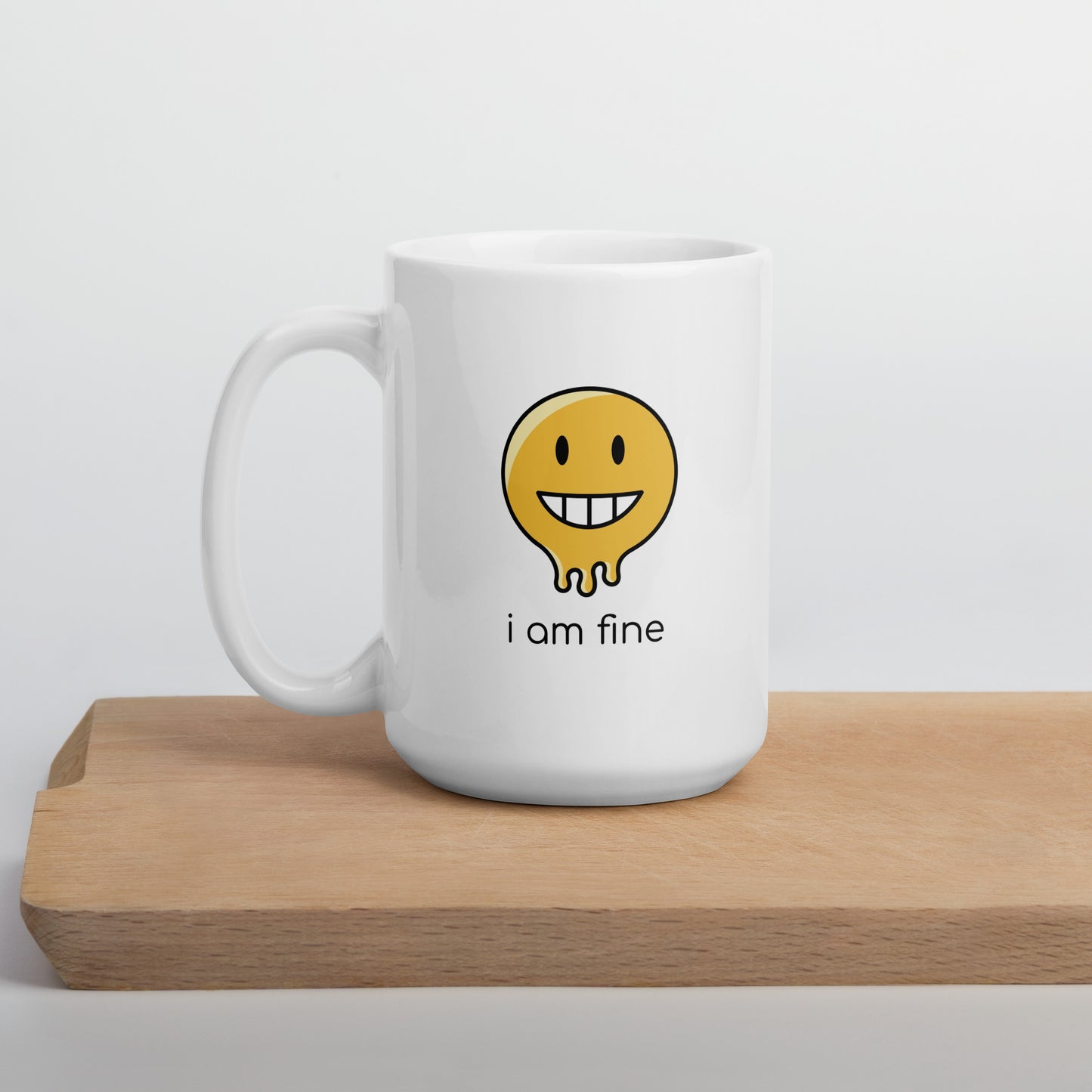 A glossy white ceramic mug with a graphic of a yellow smiley face (grinning) and melting at the bottom, accompanied by the caption 'i am fine' in small black sans serif text, positioned on a wooden cutting board against a neutral background.