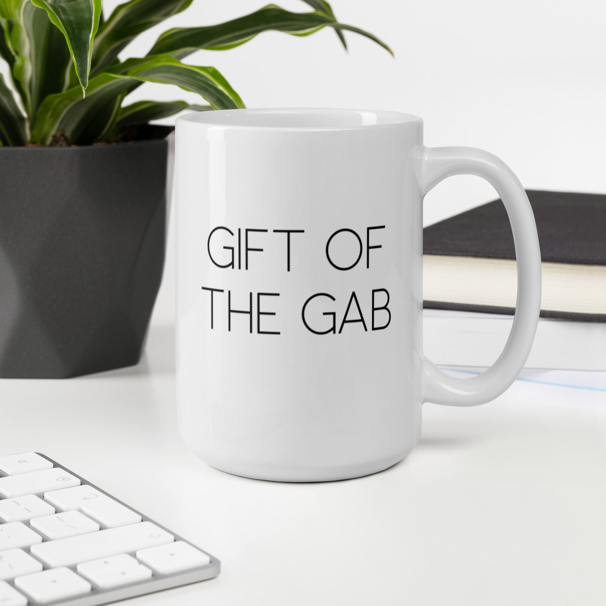 A glossy white ceramic mug with thin, all-caps black sans serif font that reads 'GIFT OF THE GAB', sits upright on a white table. In the background, there's a potted plant with downward-curving leaves, resembling a snake plant, in a dark grey pot. Accompanying the plant is a stack of books. A corner of a keyboard is partially visible in the foreground, slightly out of focus, adding a subtle hint of workspace ambiance.