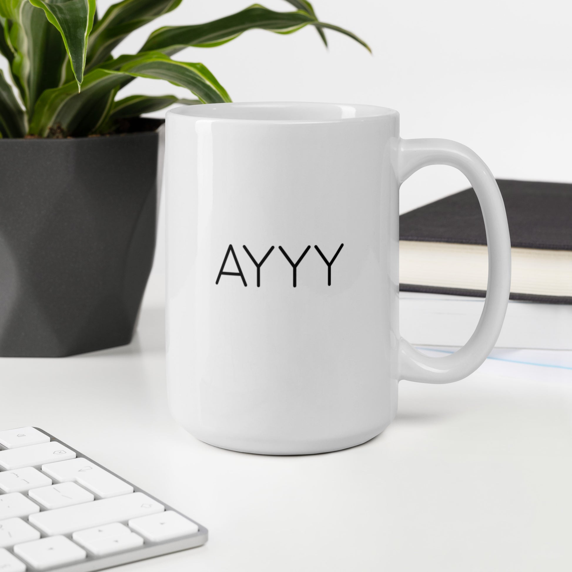 A glossy white ceramic mug with all-caps thin black text that reads 'AYYY', sits upright on a white table. In the background, there's a potted plant with downward-curving leaves, resembling a snake plant, in a dark grey pot. Accompanying the plant is a stack of books. A corner of a keyboard is partially visible in the foreground, slightly out of focus, adding a subtle hint of workspace ambiance.