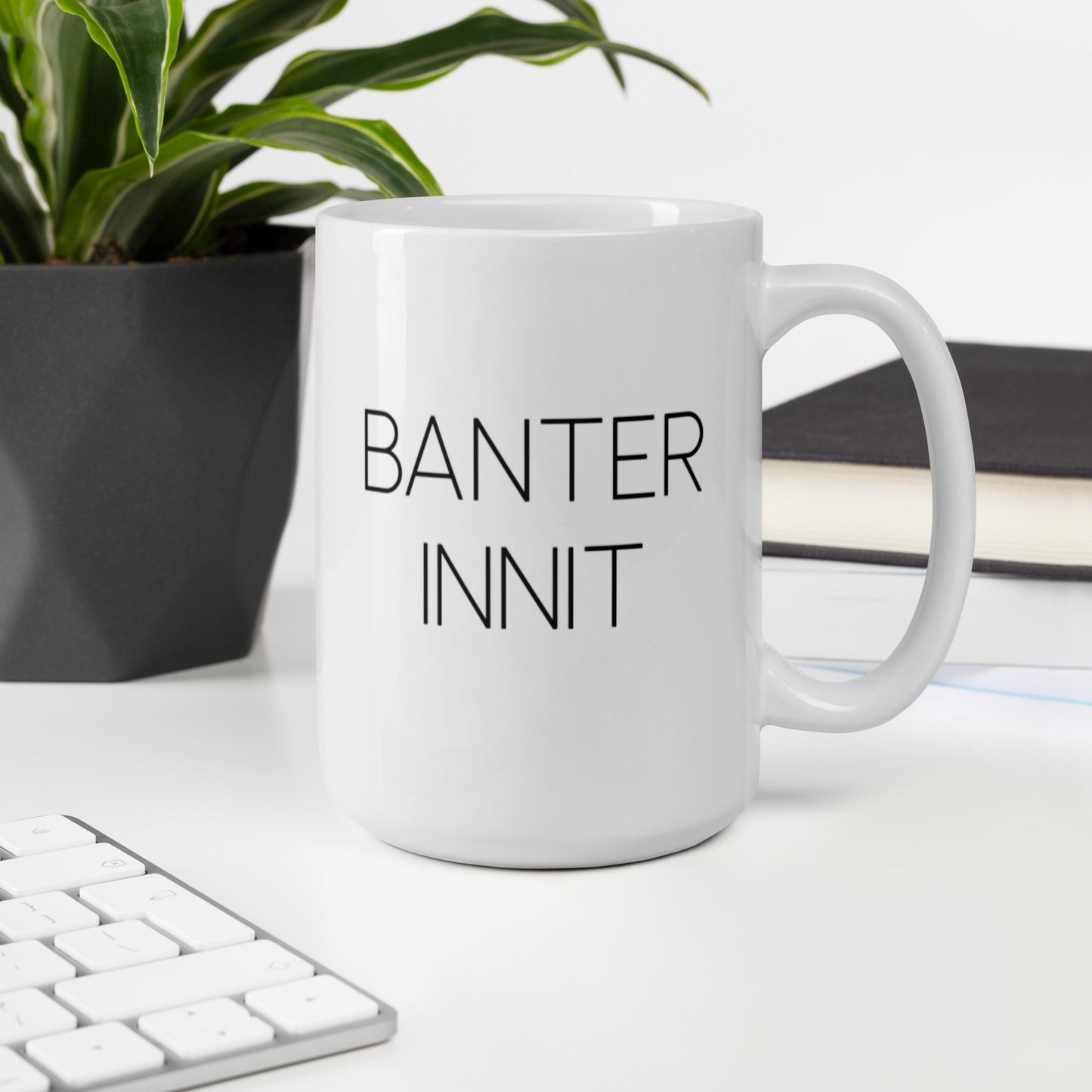 A glossy white ceramic mug with all-caps thin black text that reads 'BANTER INNIT', sits upright on a white table. In the background, there's a potted plant with downward-curving leaves, resembling a snake plant, in a dark grey pot. Accompanying the plant is a stack of books. A corner of a keyboard is partially visible in the foreground, slightly out of focus, adding a subtle hint of workspace ambiance.