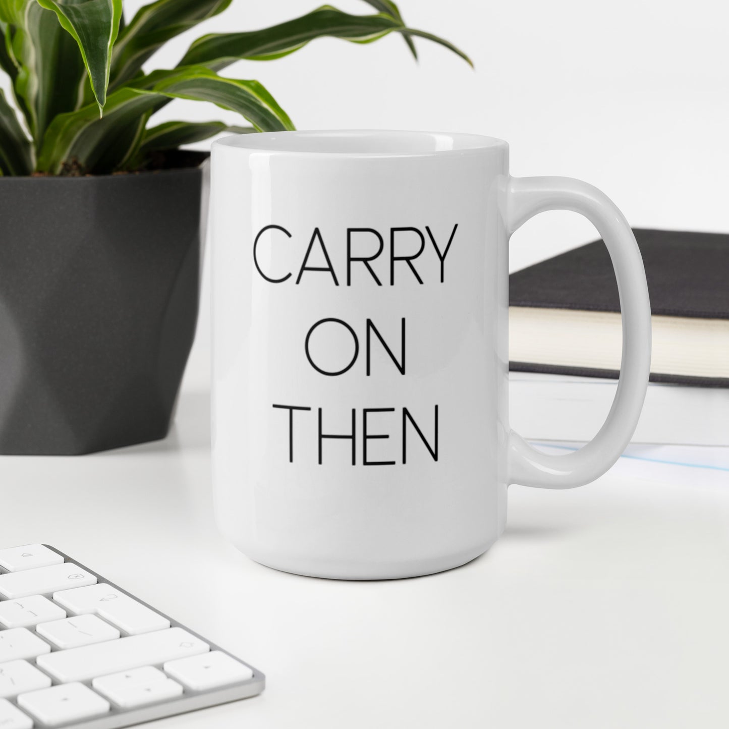 A glossy white ceramic mug with thin black all-caps text that reads 'CARRY ON THEN', sits upright on a white table. In the background, there's a potted plant with downward-curving leaves, resembling a snake plant, in a dark grey pot. Accompanying the plant is a stack of books. A corner of a keyboard is partially visible in the foreground, slightly out of focus, adding a subtle hint of workspace ambiance.