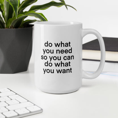 A glossy white ceramic mug with lowercase bolded black sans serif font that reads 'do what you need so you can do what you want', sits upright on a white table. In the background, there's a potted plant with downward-curving leaves, resembling a snake plant, in a dark grey pot. Accompanying the plant is a stack of books. A corner of a keyboard is partially visible in the foreground, slightly out of focus, adding a subtle hint of workspace ambiance.