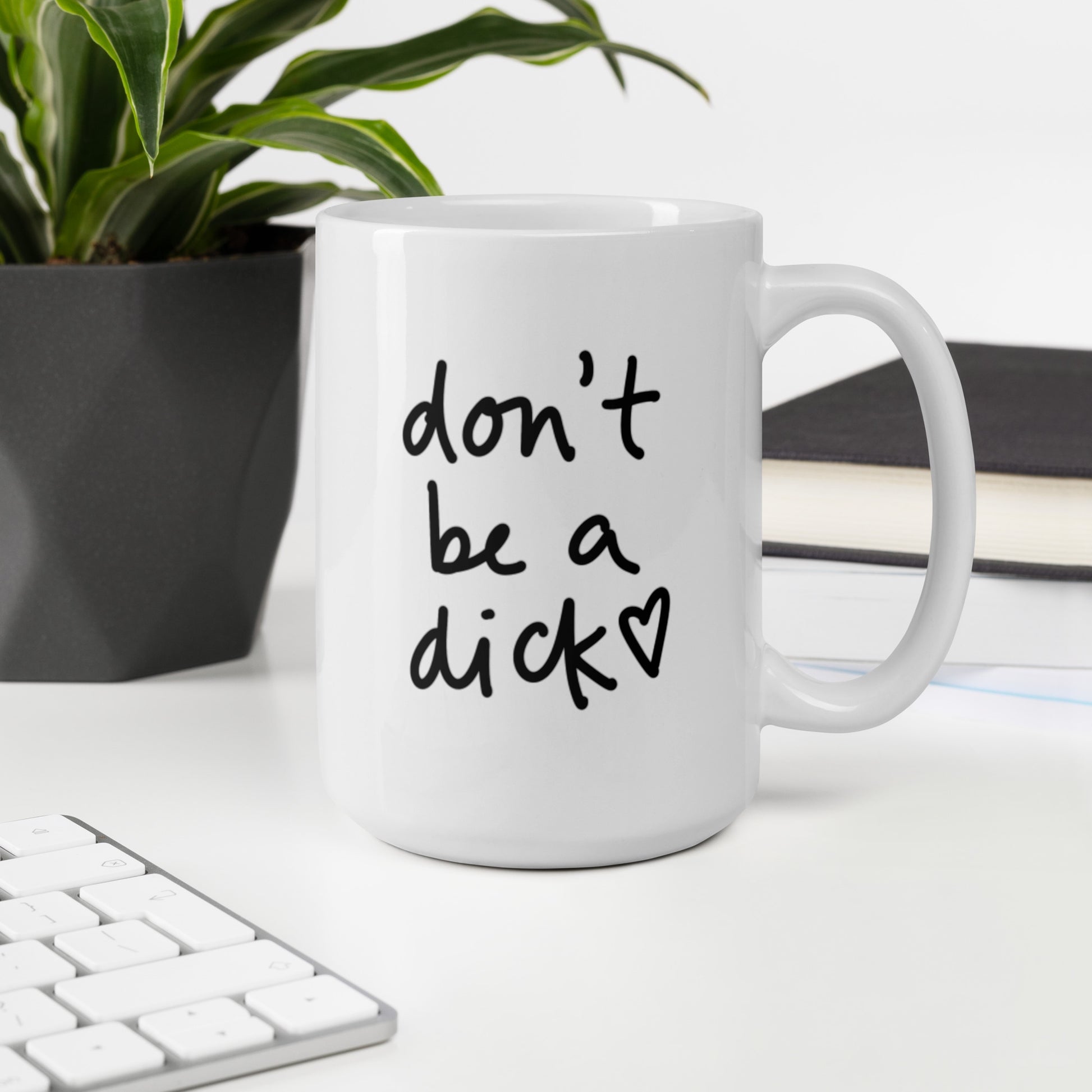 A glossy white ceramic mug with bold handwritten-style font that reads 'don't be a dick [ hand-drawn heart ]', sits upright on a white table. In the background, there's a potted plant with downward-curving leaves, resembling a snake plant, in a dark grey pot. Accompanying the plant is a stack of books. A corner of a keyboard is partially visible in the foreground, slightly out of focus, adding a subtle hint of workspace ambiance.