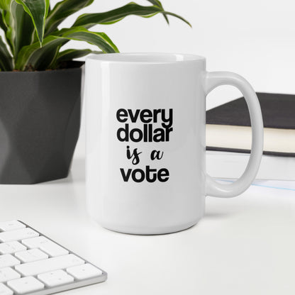 A glossy white ceramic mug with bold, lowercase black sans serif font that reads 'every dollar is a vote', with 'is a' in black handwritten script font, sits upright on a white table. In the background, there's a potted plant with downward-curving leaves, resembling a snake plant, in a dark grey pot. Accompanying the plant is a stack of books. A corner of a keyboard is partially visible in the foreground, slightly out of focus, adding a subtle hint of workspace ambiance.