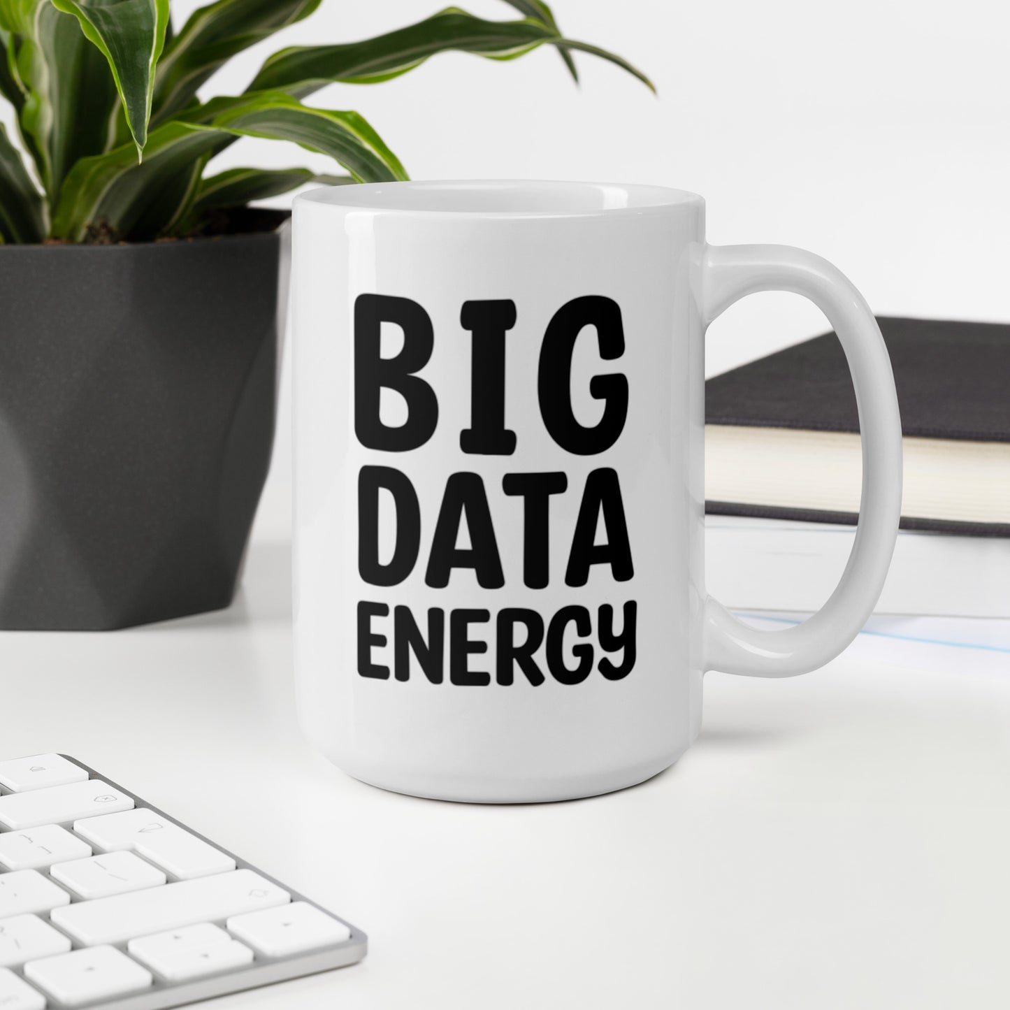 A glossy white ceramic mug with bolded black bubble text that reads 'BIG DATA ENERGY', sits upright on a white table. In the background, there's a potted plant with downward-curving leaves, resembling a snake plant, in a dark grey pot. Accompanying the plant is a stack of books. A corner of a keyboard is partially visible in the foreground, slightly out of focus, adding a subtle hint of workspace ambiance.