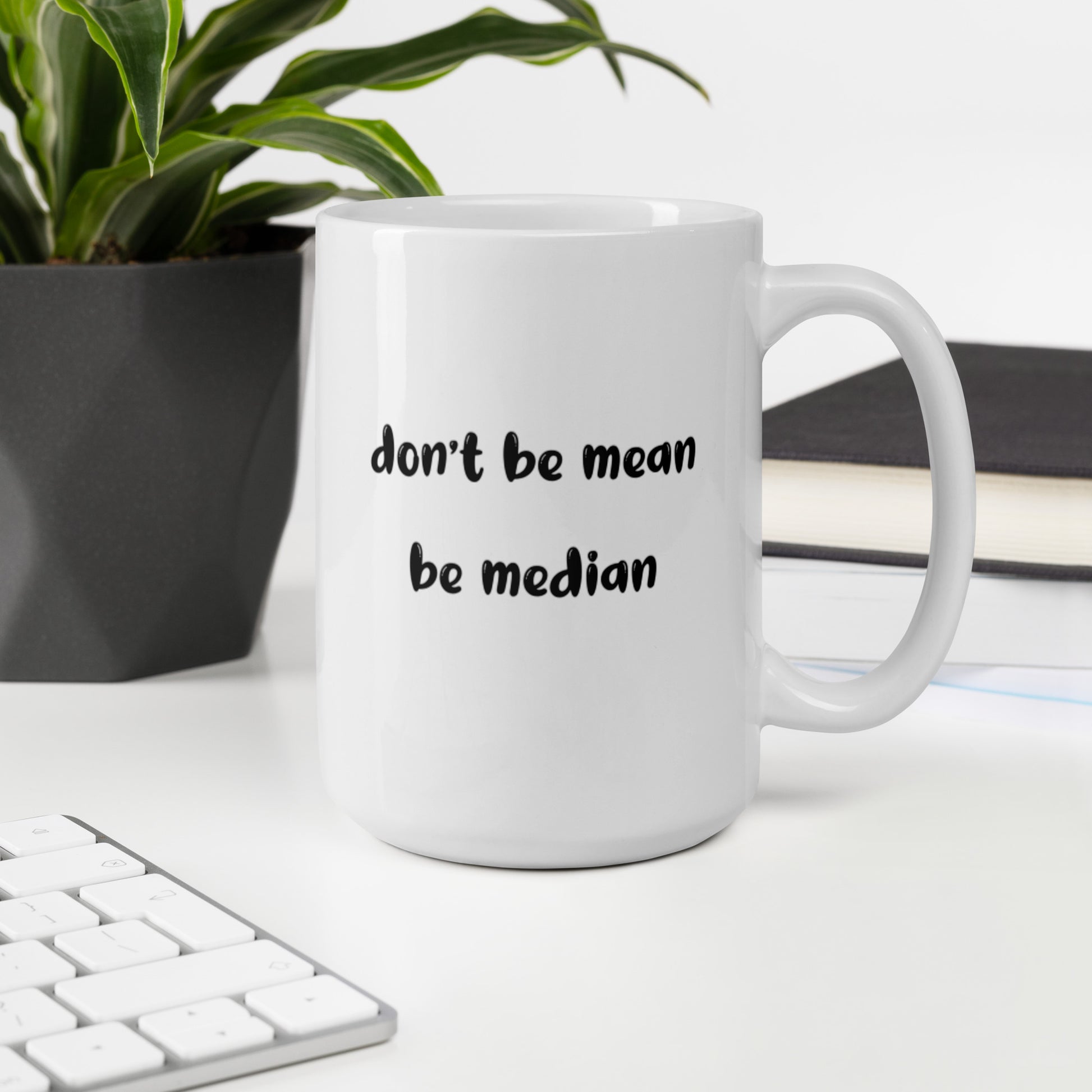 A glossy white ceramic mug with bold, lowercase black bubble font that reads 'don't be mean be median', sits upright on a white table. In the background, there's a potted plant with downward-curving leaves, resembling a snake plant, in a dark grey pot. Accompanying the plant is a stack of books. A corner of a keyboard is partially visible in the foreground, slightly out of focus, adding a subtle hint of workspace ambiance.