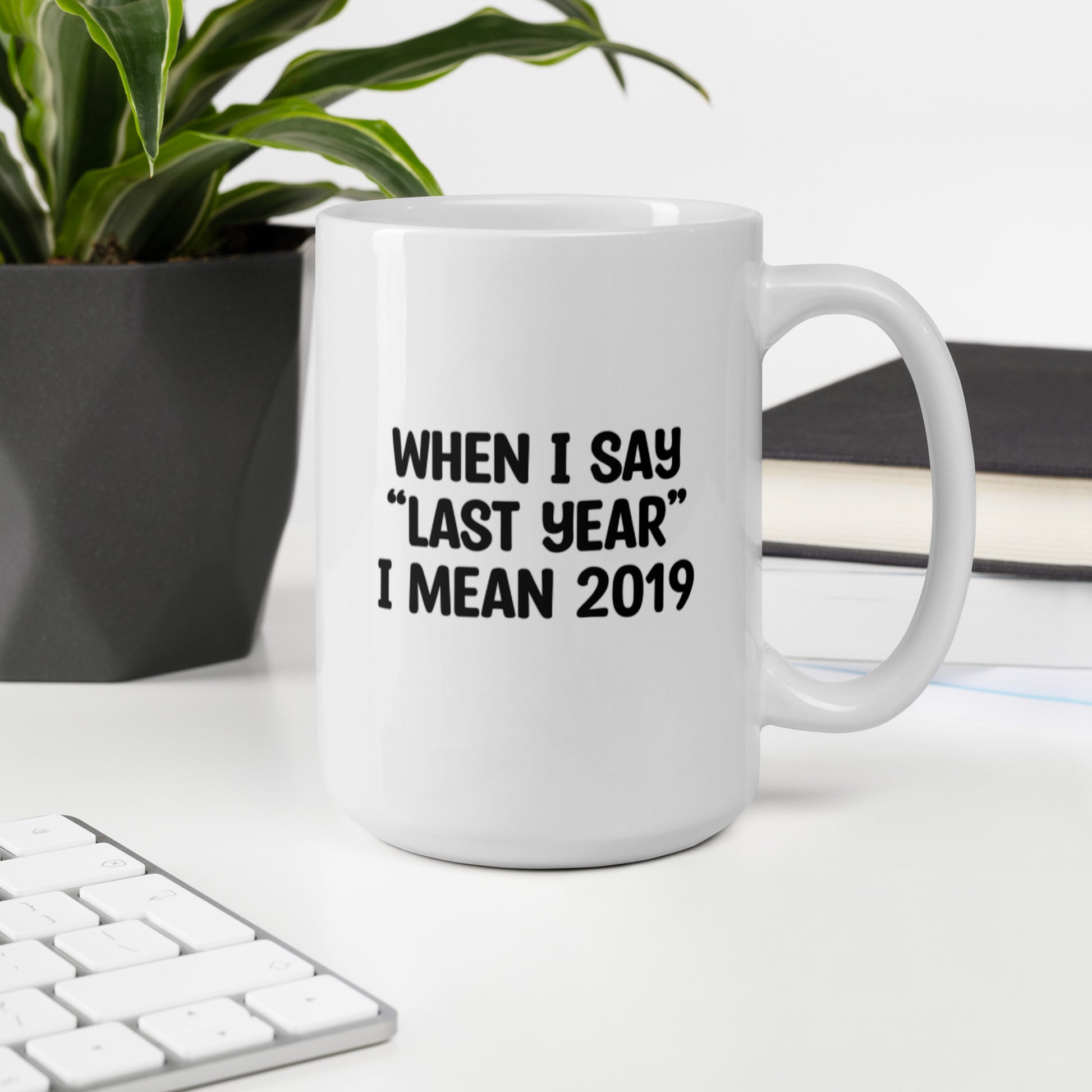 A glossy white ceramic mug with the text 'WHEN I SAY 'LAST YEAR' I MEAN 2019' in black bold uppercase font, sits upright on a white table. In the background, there's a potted plant with downward-curving leaves, resembling a snake plant, in a dark grey pot. Accompanying the plant is a stack of books. A corner of a keyboard is partially visible in the foreground, slightly out of focus, adding a subtle hint of workspace ambiance.