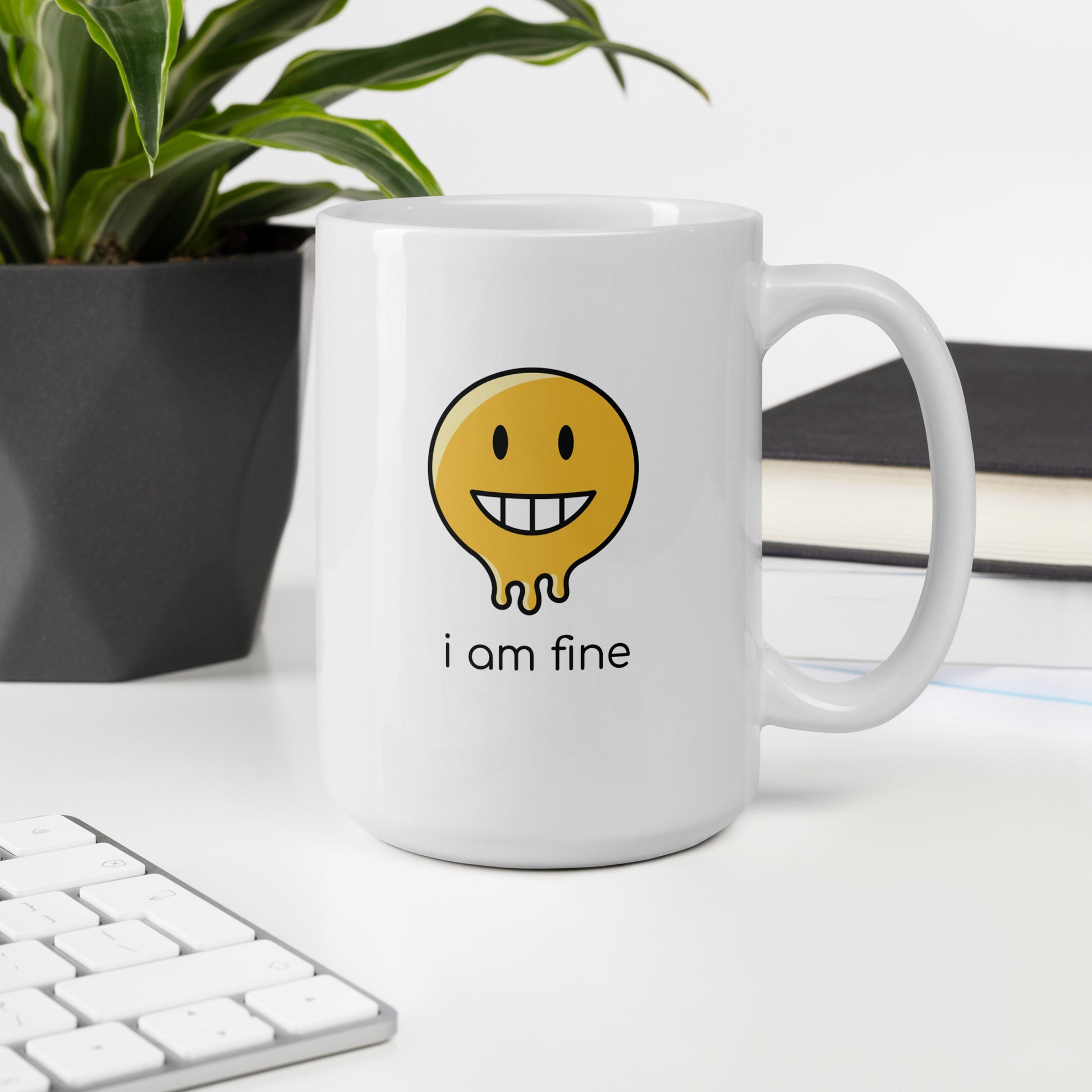 A glossy white ceramic mug with a graphic of a yellow smiley face (grinning) and melting at the bottom, accompanied by the caption 'i am fine' in small black sans serif text, sits upright on a white table. In the background, there's a potted plant with downward-curving leaves, resembling a snake plant, in a dark grey pot. Accompanying the plant is a stack of books. A corner of a keyboard is partially visible in the foreground, slightly out of focus, adding a subtle hint of workspace ambiance.